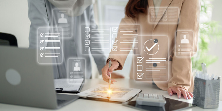 Zwei Personen stehen an einem Schreibtisch in einem Büro. Eine Person markiert mit einem Stift ein Dokument auf einem Klemmbrett. Auf dem Tisch liegen ein Laptop, ein Tablet und ein Taschenrechner. Über dem Bild sind symbolisch digitale Checklisten und Häkchen eingeblendet, die für Prüfung, Auswahl oder Bewertung stehen.<address>© stock.adobe.com - STBSTD</address>