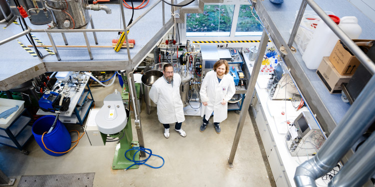 Prof. Dr. Jochen Schmid und sein Mitarbeiter Dr. Jannis Bröker stehen nebeneinander in weißen Kitteln im Biotechnikum. Die Personen sind eher im Hintergrund und von oben (von einer Galerie aus) fotografiert. Ringsherum ist Labortechnik zu sehen.<address>&copy; Uni MS - Linus Peikenkamp</address>