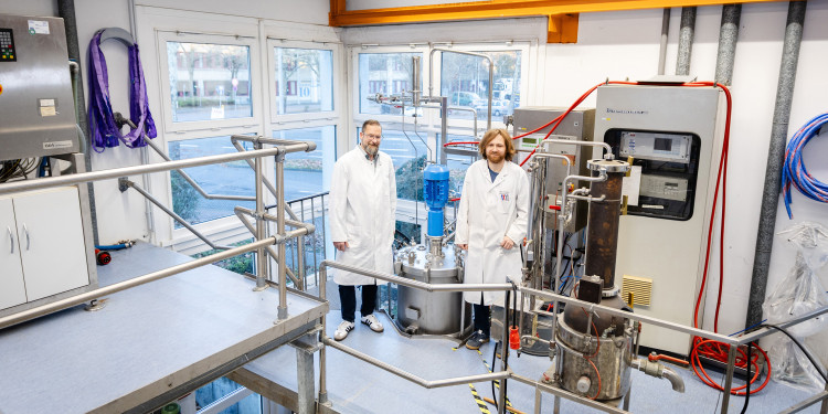 Prof. Dr. Jochen Schmid und sein Mitarbeiter Dr. Jannis Bröker stehen nebeneinander in weißen Kitteln im Biotechnikum auf einer Galerie, die die obere Hälfte des hohen Raumes begehbar macht. Die Personen sind eher im Hintergrund. Ringsherum ist Technik zu sehen, unter anderem die Spitze des größten Fermenters hinter der Galerie.<address>&copy; Uni MS - Linus Peikenkamp</address>