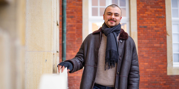 Das Bild zeigt Ferhat Kafali vor der Tür zum Südflügel des Schlosses. Er lächelt in die Kamera und hält sich am Geländer fest.<address>&copy; Uni MS - Linus Peikenkamp</address>