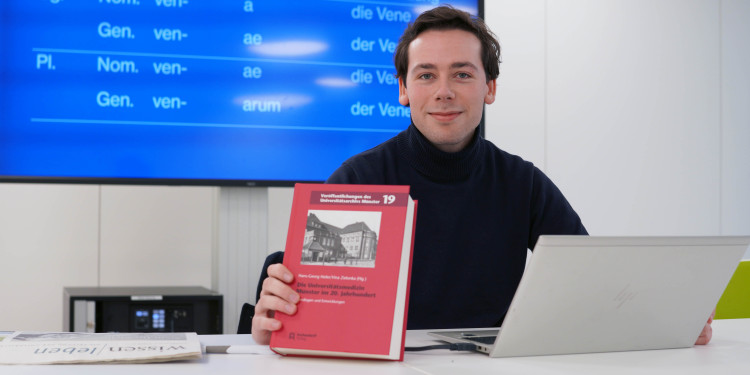 Das Bild zeigt Carl Eickmann am Pult hinter seinem Laptop. Er zeigt den Jubiläumsband zum 100-jährigen Bestehen der münsterschen Universitätsmedizin in die Kamera. Hinter ihm ist ein großer Bildschirm mit einer Präsentation und lateinischen Vokabeln zu sehen.<address>&copy; Uni MS - Julia Harth</address>