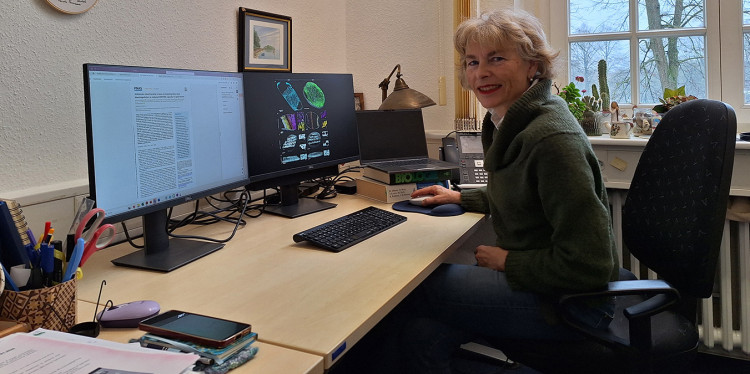 Prof. Dr. Karin Busch at her desk in front of two monitors. She turns sideways to face the photographer. The right-hand screen shows images of mitochondria, while the left-hand screen displays one of her current papers.<address>&copy; Uni MS - Christina Hoppenbrock</address>