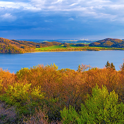 Blick auf den Laacher See in der östlichen Vulkaneifel (Panorama). Der See ist umgeben von hügeliger Landschaft mit herbstlich verfärbten Bäumen, der Himmel ist blau mit Wolken.<address>&copy; mojolo - stock.adobe.com</address>