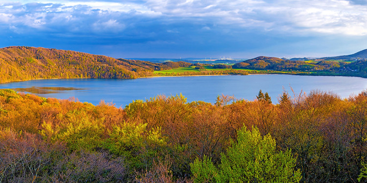 Blick auf den Laacher See in der östlichen Vulkaneifel (Panorama). Der See ist umgeben von hügeliger Landschaft mit herbstlich verfärbten Bäumen, der Himmel ist blau mit Wolken.<address>&copy; mojolo - stock.adobe.com</address>