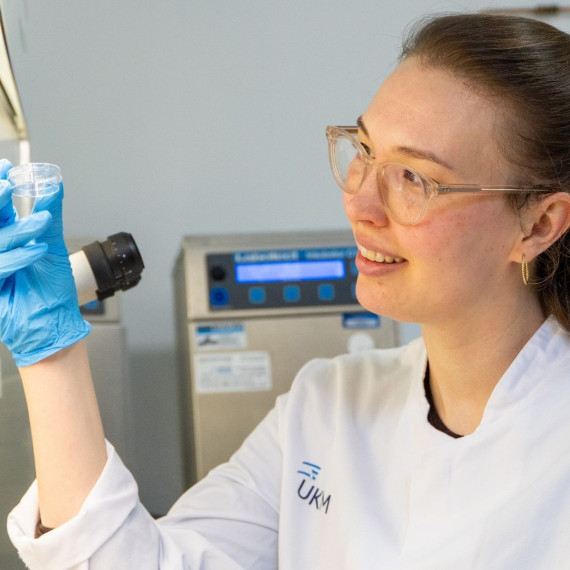 Eine Wissenschaftlerin hält eine Proben­schale und untersucht sie im medizinischen Labor an einem Mikroskop.<address>&copy; Uni MS - Linus Peikenkamp</address>