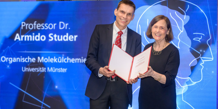 Zwei Personen stehen vor einer blau beleuchteten Wand, auf der der Name des Preisträgers (Armido Studer) geschrieben steht. Armido Studer steht links, die Präsidentin der DFG rechts, beide lachen.<address>&copy; DFG / David Ausserhofer</address>