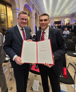 Zwei Männer stehen in einem festlich beleuchteten Saal mit hohen Bögen und halten gemeinsam eine geöffnete Urkunde in einer roten Mappe in die Kamera. Der Mann rechts (Armido Studer) lächelt, trägt einen dunklen Anzug mit roter Krawatte; links steht Rektor Johannes Wessels im Anzug. Im Hintergrund sitzen und stehen weitere Gäste der Veranstaltung.<address>&copy; Uni MS - Norbert Robers</address>