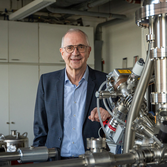 Prof. Dr. Harald Fuchs steht hinter einem - zum Teil sichtbaren - Rastersondenmikroskop in einem Labor und blickt in die Kamera (Porträtaufnahme).<address>&copy; Uni MS - Linus Peikenkamp</address>