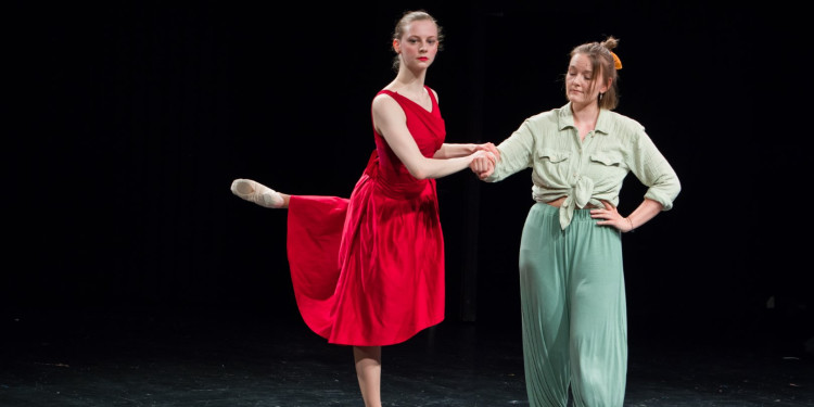 Zwei Frauen stehen auf der Bühne des Stadttheaters Münster vor schwarzem Hintergrund. Die linke Frau hat ein rotes Kleid an, tanzt Ballett und hält sich am Arm der anderen Frau in grüner Kleidung fest.<address>&copy; Ralf Emmerich</address>