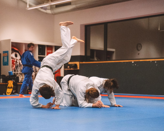 Das Bild zeigt drei Judo-Sportlerinnen in weißen Anzügen bei einer Übung auf der Matte - die vordere Person macht dabei eine Art Handstand.<address>&copy; Hochschulsport</address>