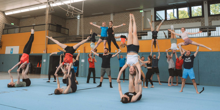 Das Bild zeigt etwa 20 Sportlerinnen und Sportler in einer Turnhalle, die Akrobatikübungen und Hebefiguren machen.<address>&copy; Hochschulsport</address>