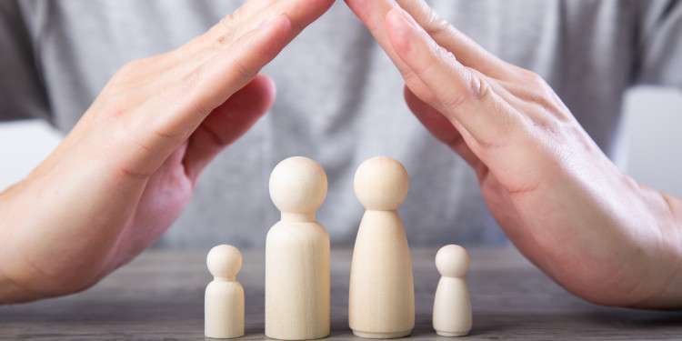 Man sieht zwei Hände, die zu einem Dach geformt sind. Darunter stehen vier Holzfiguren, die eine Familie darstellen.<address>&copy; stock.adobe.de - chee siong teh</address>