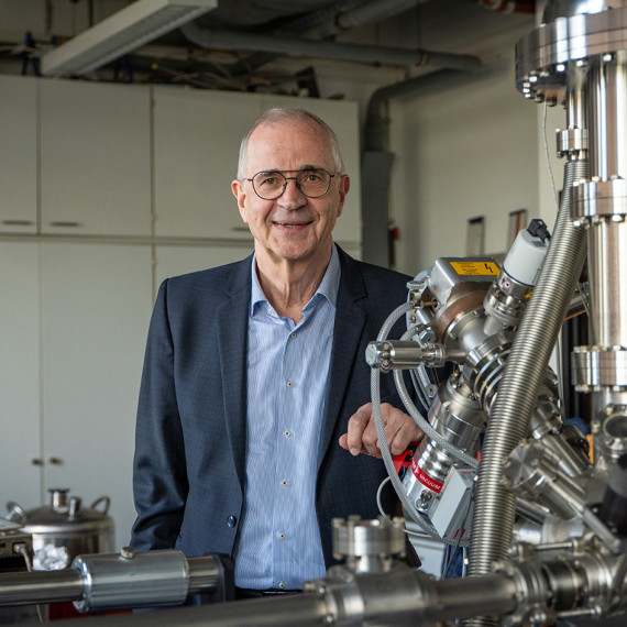 Prof. Dr Harald Fuchs is standing behind a scanning probe microscope – part of which is visible – in a laboratory and looking into the camera (portrait shot).<address>&copy; Uni MS - Linus Peikenkamp</address>