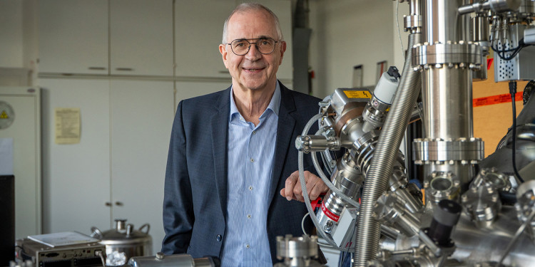 Prof. Dr Harald Fuchs is standing behind a scanning probe microscope – part of which is visible – in a laboratory and looking into the camera (portrait shot).<address>&copy; Uni MS - Linus Peikenkamp</address>