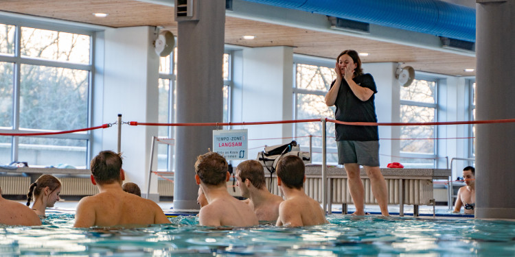 Das Bild zeigt ein Schwimmbecken, in dem sich mehrere Personen befinden. Im Hintergrund steht am Beckenrand die Sportdozentin Maike Elbracht. Sie gibt den Studierenden im Wasser Anweisungen.<address>&copy; Uni MS - Linus Peikenkamp</address>