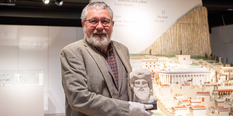 Dr. Helge Nieswandt präsentiert den Gipsabguss eines griechischen Gottes. Er steht im Museum und trägt Handschuhe. Im Hintergrund ist das Modell einer antiken Stadt zu sehen.<address>&copy; Uni MS - Linus Peikenkamp</address>
