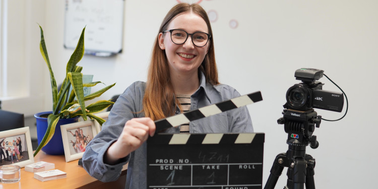 Luise Hecht steht in ihrem Büro vor einem Sideboard und lächelt in die Kamera. Sie hält eine Filmklappe in der Hand. Neben ihr steht eine Kamera auf einem Stativ.<address>&copy; Uni MS - Julia Harth</address>