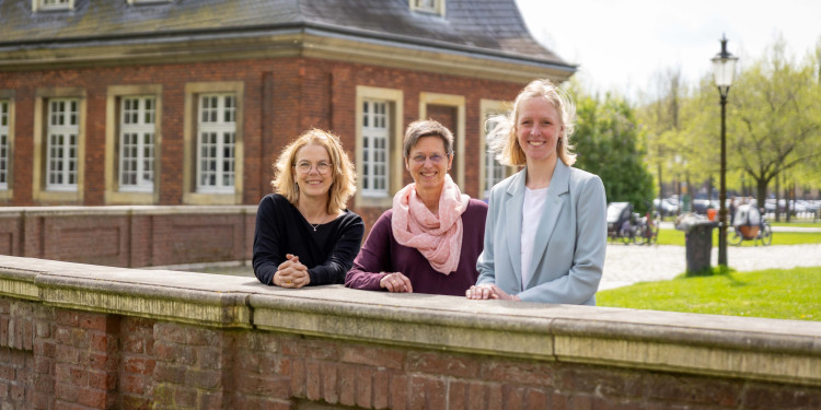 Prof. Dr. Maike Tietjens, Prof. Dr. Stefanie van Ophuysen und Projektreferentin Dr. Helen Jäckel (von links) stehen hinter einer Mauer, lehnen sich auf diese und schauen in die Kamera. Hinter ihnen ist das Kavaliershäuschen vor dem Schloss zu sehen.<address>&copy; Uni MS - Linus Peikenkamp</address>