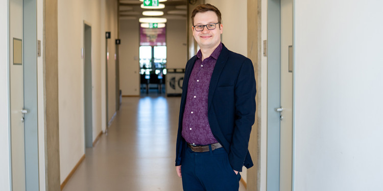 Prof. Dr. Robert Hein steht rechts im Bild, dem Betrachter zugewandt (seitlich, zur Kamera gedreht). Er steht in einem nach hinten verlaufenden Flur mit Türen rechts und links; die Tür im Bildhintergrund vor Kopf des Flures ist geöffnet. Im Inneren des Raumes ist unscharf ein Besprechungstisch zu sehen, hinter dem Tisch sind Fenster.<address>&copy; Uni MS - Linus Peikenkamp</address>