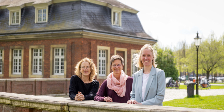 Prof. Dr Maike Tietjens, Prof. Dr Stefanie van Ophuysen and Project Consultant Dr Helen Jäckel (from left) are standing behind a wall, leaning against it and looking at the camera. Behind them, the Kavaliershäuschen can be seen.<address>&copy; University of Münster - Linus Peikenkamp</address>