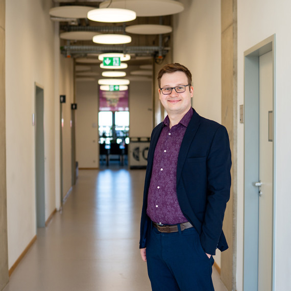 Prof. Robert Hein is standing on the right of the picture, facing the viewer (from the side, turned towards the camera). He is standing in a corridor leading away from the camera, with doors on the right and left; the door at the far end of the corridor, visible in the background, is open. A conference table can be seen out of focus inside the room, with windows behind it.<address>&copy; Uni MS - Linus Peikenkamp</address>
