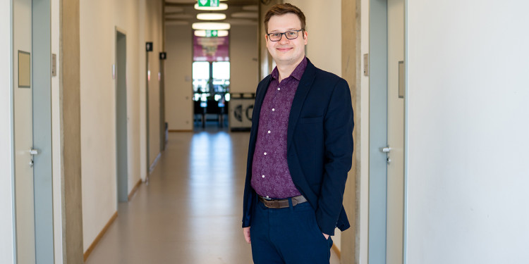 Prof. Robert Hein is standing on the right of the picture, facing the viewer (from the side, turned towards the camera). He is standing in a corridor leading away from the camera, with doors on the right and left; the door at the far end of the corridor, visible in the background, is open. A conference table can be seen out of focus inside the room, with windows behind it.<address>&copy; Uni MS - Linus Peikenkamp</address>