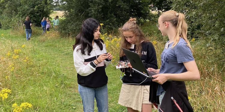 Drei Teilnehmerinnen eines früheren Workshops zum Thema „Nature meets IT“ stehen mit einem Laptop und erkennbaren Sensoren auf einer Wiese. Im Hintergrund stehen Bäume und man sieht vier weitere Teilnehmerinnen..<address>&copy; MExLab ExperiMINTe</address>