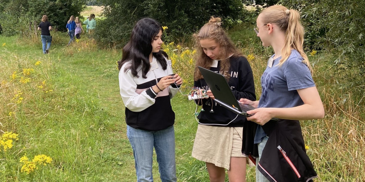 Drei Teilnehmerinnen eines früheren Workshops zum Thema „Nature meets IT“ stehen mit einem Laptop und erkennbaren Sensoren auf einer Wiese. Im Hintergrund stehen Bäume und man sieht vier weitere Teilnehmerinnen..<address>&copy; MExLab ExperiMINTe</address>