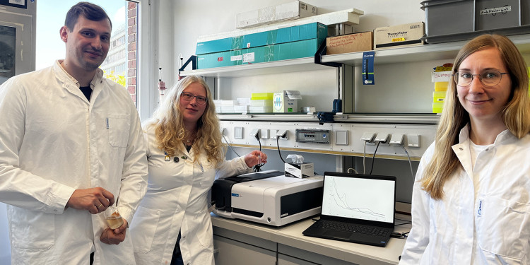 Prof. Julia Kurth (right) with doctoral students Lukas Hofmann and Jasmin Bernhardt. The three are standing in a laboratory in front of a workbench and are wearing white lab coats. All three are facing the photographer and looking into the camera.<address>&copy; AG Kurth - Zeynep Doğru</address>