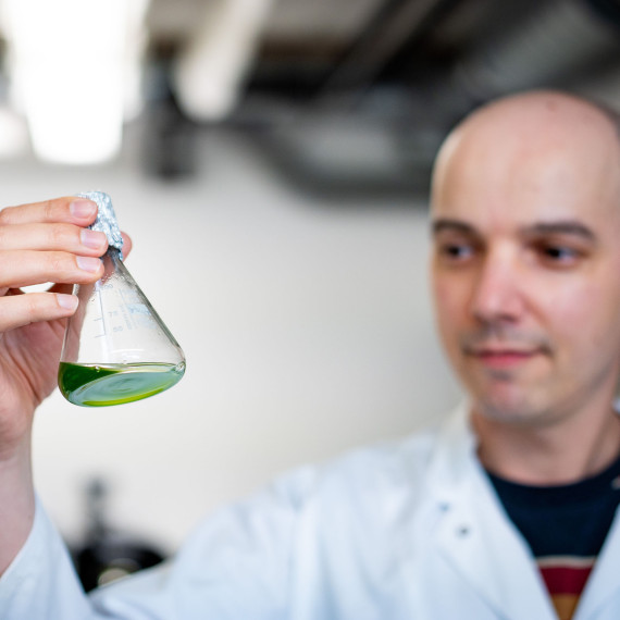 A man is standing in a laboratory (wearing a white coat; the frame is cropped at chest height). He is slightly out of focus. In the foreground, in sharp focus, is a glass flask containing single-celled green algae, which he is holding up and examining.<address>&copy; Uni MS - Linus Peikenkamp</address>