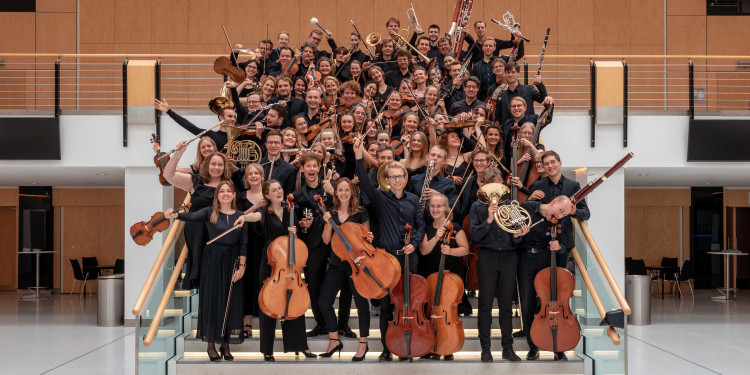 Das Bild zeigt das Ensemble in der Kongresshalle der Halle Münsterland. Etwa vier Dutzend Musikerinnen und Musiker stehen in dunkler Abendgarderobe eng nebeneinander auf einer Treppe und, schauen fröhlich in die Kamera und präsentieren ihre Instrumente.<address>&copy; Adam Karas</address>
