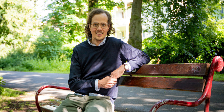 Das Bild zeigt Colin Schulz auf einer Bank sitzend. Im Hintergrund sind die Promenade und Bäume zu sehen.<address>&copy; Uni MS – Linus Peikenkamp</address>