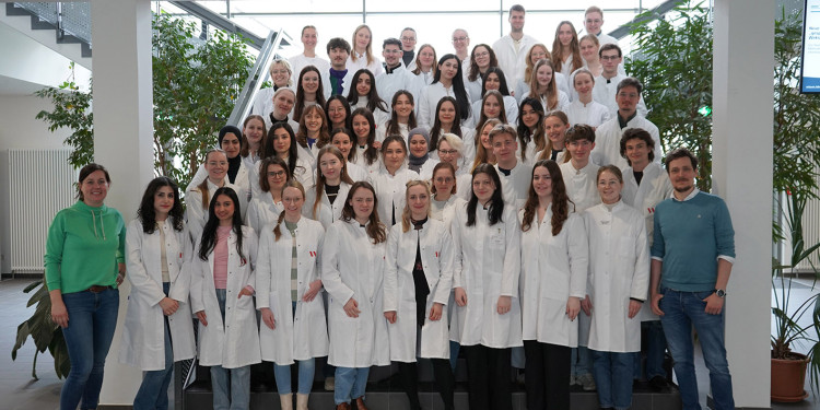 Eine Gruppe von Studierenden in weißen Laborkittel. Links steht eine Frau in grünem Pullover, rechts ein Mann mit blauem Pullover.<address>&copy; PharMSchool - Dr. Fabian Herrmann</address>