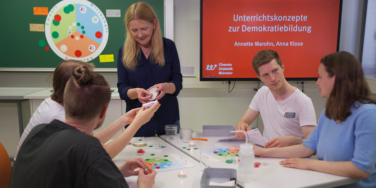 Eine Szene in einem Seminar: Vier Studierende sitzen um einen Tisch mit Unterrichtsmaterialien/Experimentierzubehör. Die Professorin steht hinter dem Tisch und ist im Gespräch mit den Studierenden und demonstriert etwas. Im Hintergrund auf einem Monitor steht als weißer Schriftzug auf rotem Grund: "Unterrichtskonzepte zur Demokratiebildung" (Annette Marohn, Anna Klose; Chemie Didaktik Münster)<address>&copy; Annette Marohn</address>