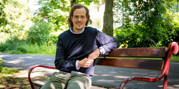 The picture shows Colin Schulz sitting on a bench. The promenade and trees can be seen in the background.<address>&copy; Uni MS – Linus Peikenkamp</address>