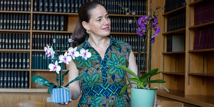A woman standing in front of a bookcase. She is holding a flowerpot containing an orchid in each hand and is looking at the plant in her left hand (on the viewer’s right). The plant in her right hand is made of Lego.<address>&copy; Uni MS - Linus Peikenkamp</address>