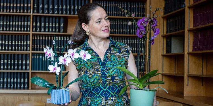 Eine Frau vor einer Bücherwand. Sie hat in jeder Hand einen Blumentopf mit einer Orchidee und blickt auf die Pflanze in ihrer linken Hand (von Betrachter aus rechts). Die Pflanze in ihrer rechten Hand ist aus Lego.<address>&copy; Uni MS - Linus Peikenkamp</address>