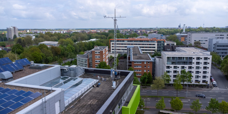 Drohnenaufnahme des GEO1-Gebäudes, auf dem an einer Seite der Messturm mehrere Meter in die Höhe ragt. Dahinter ist die Silhouette der Stadt Münster zu sehen.<address>&copy; Uni MS - Johannes Wulf</address>