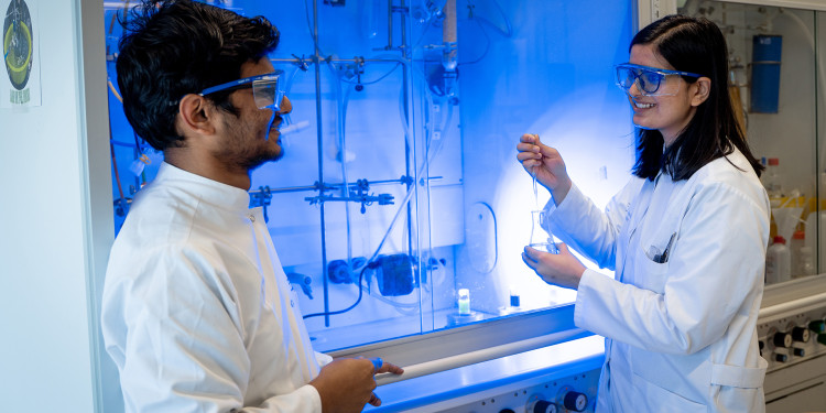 Ein Mann (links) und eine Frau (rechts) vor einem Laborabzug. Das Licht unter dem Abzug schimmert bläulich, die Frau hat eine Pipette und ein Glasgefäß in der Hand. Beide tragen weiße Laborkittel und eine Schutzbrille.<address>&copy; Uni MS - Linus Peikenkamp</address>