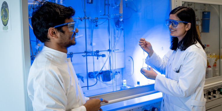 A man (left) and a woman (right) standing in front of a fume cupboard in a chemistry laboratory. The light beneath the fume cupboard has a bluish glow; the woman is holding a pipette and a glass vessel. Both are wearing white lab coats and safety glasses.<address>&copy; Uni MS - Linus Peikenkamp</address>