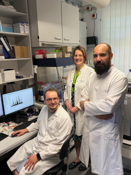 Three people wearing white lab coats. A man (on the left) is sitting at a desk in a laboratory or office; there is a monitor to his left behind him. A woman is standing to his right behind him, and another man is standing to his right beside him.<address>&copy; AG Finkemeier</address>
