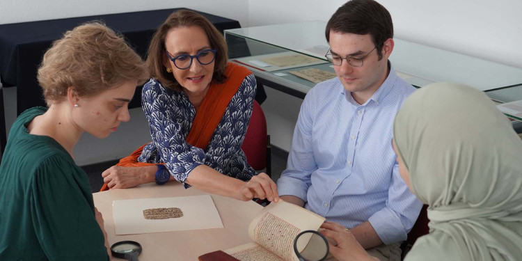Drei Studierende sitzen mit ihrer Seminarleiterin um einen Tisch herum. Darauf liegt eine alte Handschrift, ein Brieffragment und eine große Lupe. Die Studentin vorne rechts im Bild (von hinten fotografiert) hält eine zweite Lupe in der Hand.<address>&copy; Uni MS - Julia Harth</address>