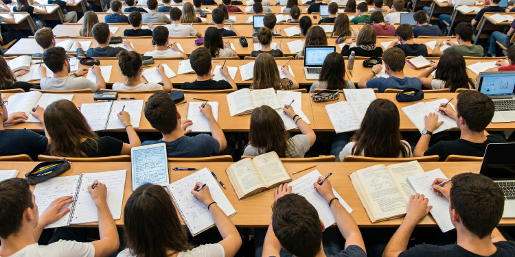 Studierende sitzen in einem Hörsaal und schreiben eine Klausur. Man sieht sie von hinten, wie sie auf ihre Zettel schreiben.<address>&copy; stock.adobe.com - Bohdan</address>