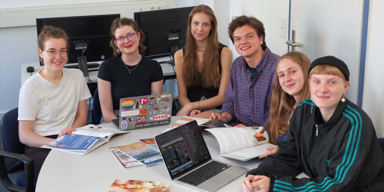 Sieben Studierende sitzen in der Lehrredaktion um einen Tisch herum und schauen in die Kamera. Auf dem Tisch steht ein Laptop und es liegen mehrere Exemplare des Magazins auf dem Tisch.<address>&copy; Uni MS - Julia Harth</address>