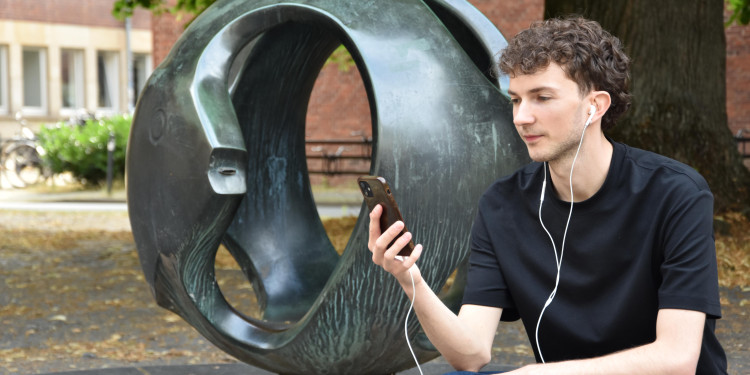 Das Bild zeigt Luca Scholz. Er hockt vor dem Fischkugelbrunnen, hält das Handy in der Hand und hat Kopfhörer in den Ohren.<address>&copy; Uni MS - Julia Harth</address>