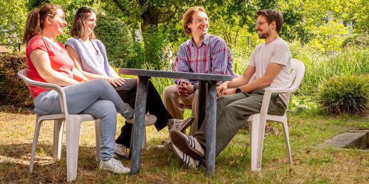 Vier Studierende sitzen an einem Tisch im Freien auf einer Wiese. Im Hintergrund steht ein Baum, dahinter sind Hecken zu erkennen.<address>&copy; Uni MS – Linus Peikenkamp</address>
