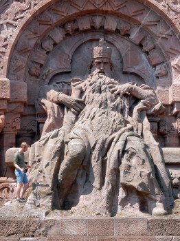 Ein Mann in grünem Shirt und blauer Shorts steht klein wirkend links vor einer überlebensgroßen steinernen Sitzstatue des bärtigen Kaisers Friedrich Barbarossa mit Krone. Die aus rötlichem Sandstein gearbeitete Figur ist Teil des Kyffhäuserdenkmals und wird von einem reich verzierten Rundbogen gerahmt.<address>&copy; Stephan Schretzenmayr/Gommern</address>