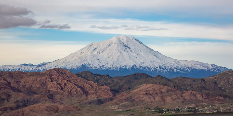 Der schneebedeckte Gipfel des Ararat erhebt sich als markanter, kegelförmiger Berg über eine weite Landschaft. Die obere Hälfte des Bergs ist von einer weißen Eiskappe bedeckt, darunter liegen kahle, bräunliche Hänge. Über dem Gipfel spannt sich ein klarer Himmel.<address>&copy; stock.adobe.com - Esin Deniz</address>