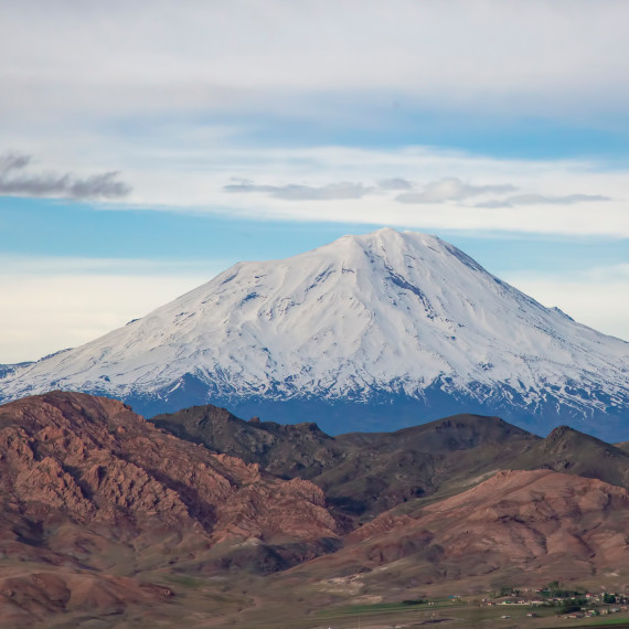 The snow-capped summit of Mount Ararat rises as a striking, conical mountain above a vast landscape. The upper half of the mountain is covered by a white ice cap, beneath which lie bare, brownish slopes. A clear sky stretches out above the summit.<address>&copy; stock.adobe.com - Esin Deniz</address>