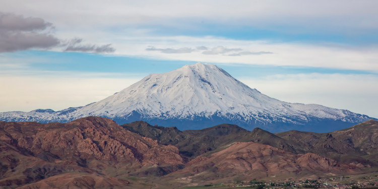 The snow-capped summit of Mount Ararat rises as a striking, conical mountain above a vast landscape. The upper half of the mountain is covered by a white ice cap, beneath which lie bare, brownish slopes. A clear sky stretches out above the summit.<address>&copy; stock.adobe.com - Esin Deniz</address>